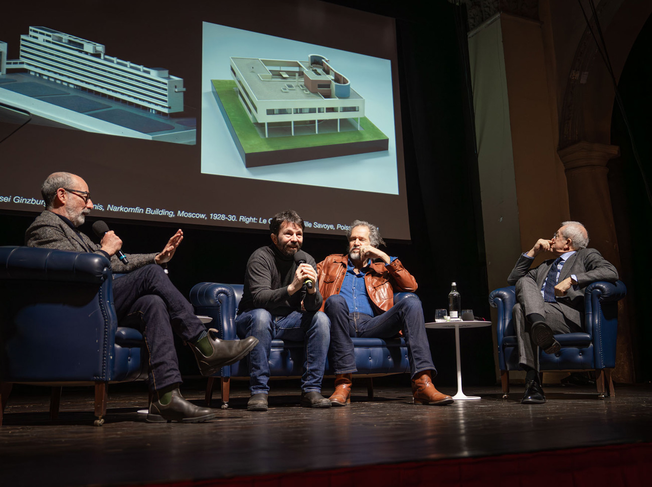 Dibattito sul palco con proiezione di modelli architettonici sullo sfondo.