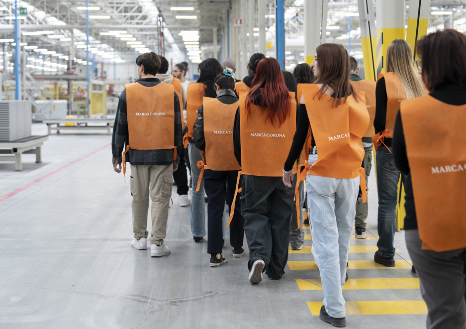 A group of students visiting the Marca Corona production department, wearing orange bibs and following a marked route inside the factory.