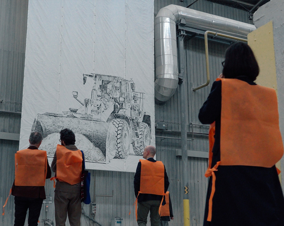 Participants wearing orange bibs observe a large technical drawing displayed on canvas inside a factory.
