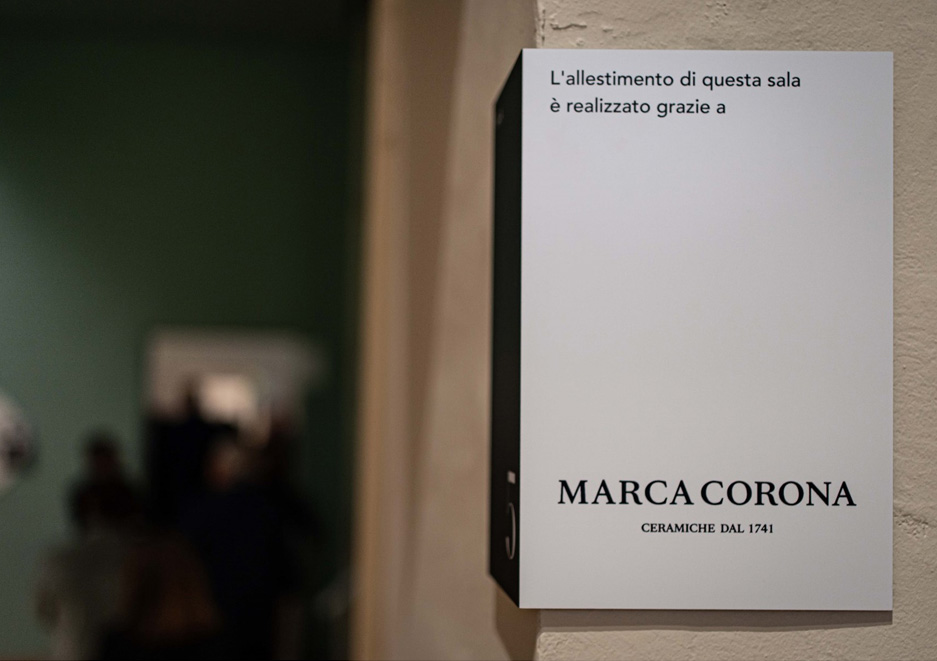 Due donne osservano un pannello espositivo illuminato, durante una mostra culturale ospitata in uno spazio storico.
