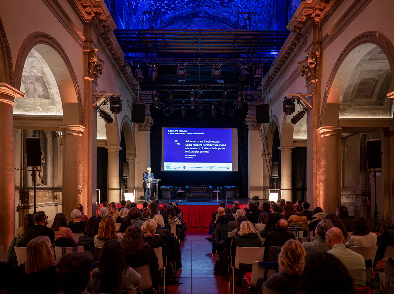 Conferenza in sala storica con relatore sul palco e pubblico seduto.