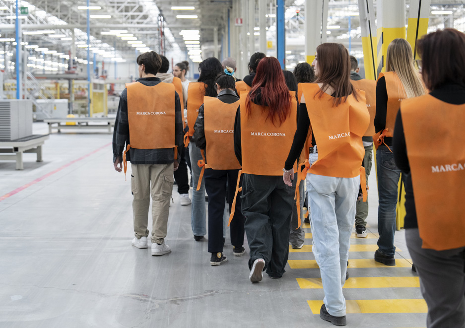 Gruppo di studenti in visita al reparto produttivo di Marca Corona, con pettorine arancioni e percorso tracciato all’interno dello stabilimento.