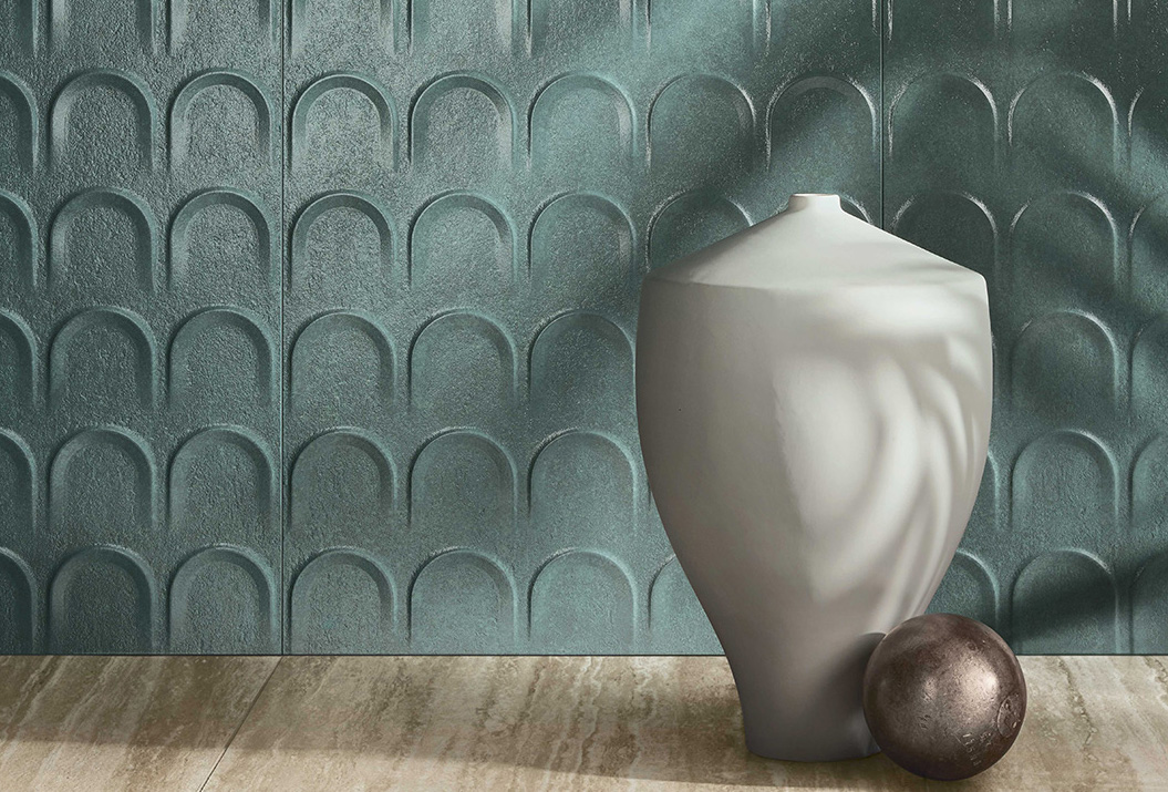 Wall covered in green ceramic tiles with an arched texture and textured surface, next to a light-coloured sculptural vase and metal sphere.