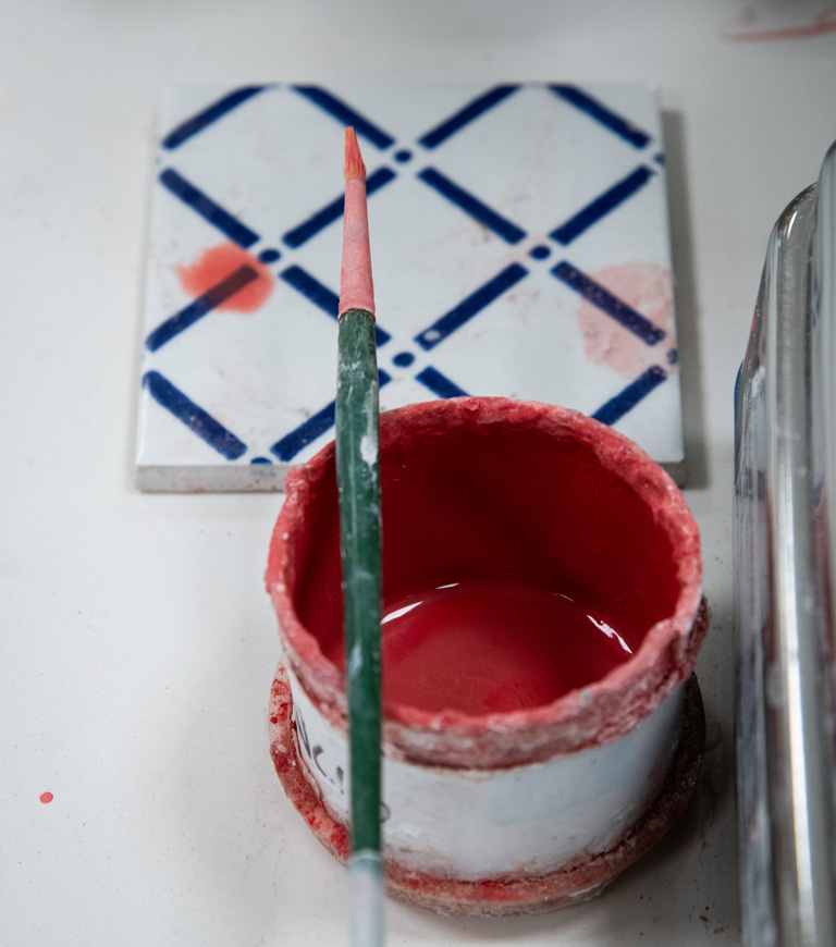Paintbrush dipped in red glaze beside a decorated ceramic tile in the ceramics laboratory.