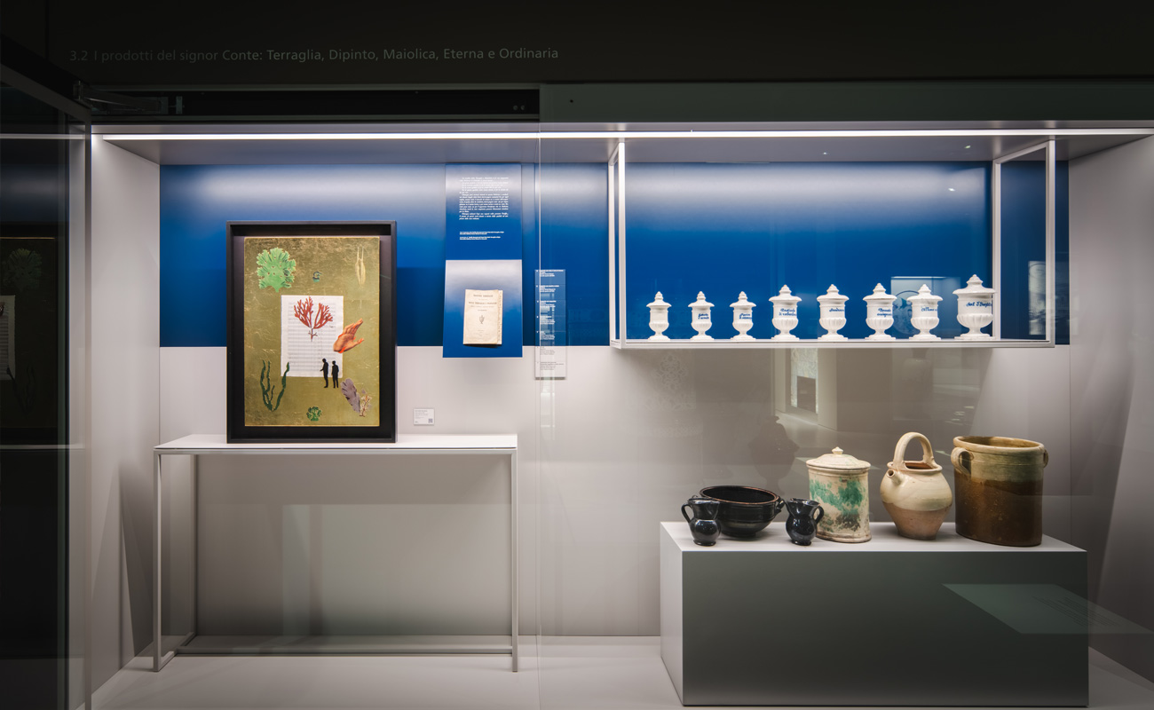 Vitrina de museo con recipientes y vasijas de cerámica, una obra enmarcada y paneles informativos sobre fondo azul.