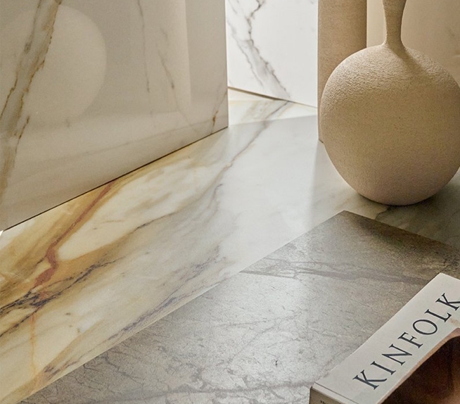 Detailansicht von Keramikoberflächen in Marmoroptik mit goldenen und braunen Maserungen, kombiniert mit materiellen Objekten in neutralen Farbtönen.