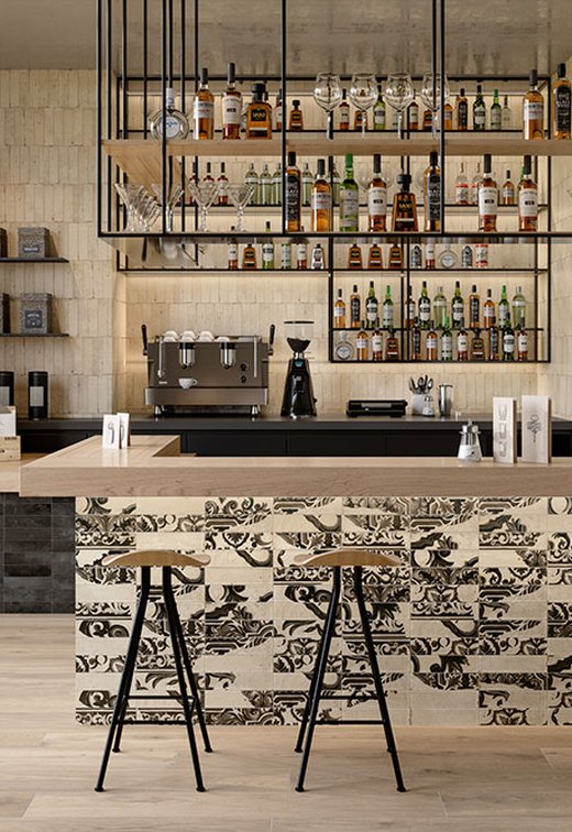 Bancone bar rivestito in gres effetto maiolica nero su fondo beige, pareti ceramiche materiche e arredi in legno chiaro e metallo.