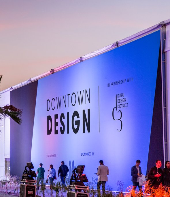 Vista esterna serale di un evento di design con padiglione illuminato e skyline urbano sullo sfondo.