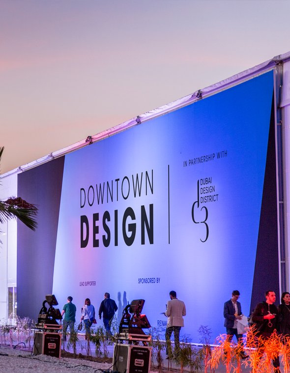 Evening exterior view of a design event with an illuminated pavilion and city skyline in the background.