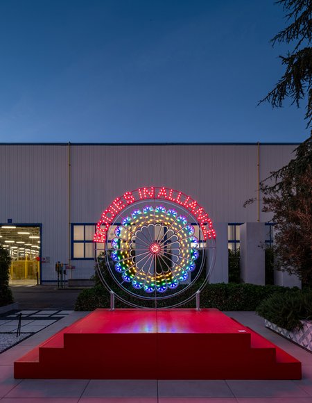 Instalación luminosa circular multicolor con el texto «BOUQUETS IN ALLIANO» frente a un edificio industrial al anochecer.