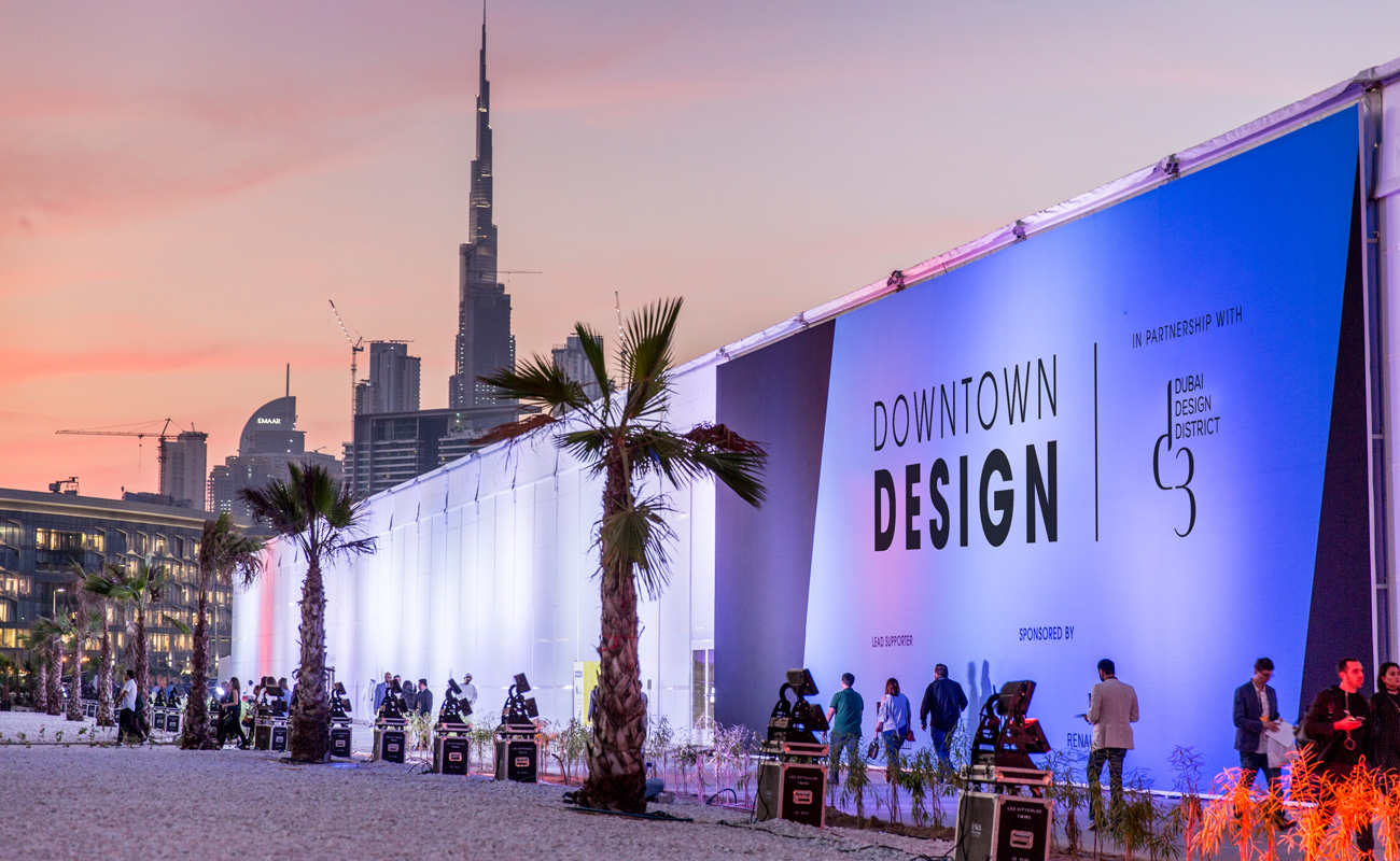 Evening exterior view of a design event with an illuminated pavilion and city skyline in the background.